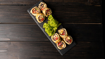 Custom sushi roll in tempura with nori, fresh salmon, tuna, avocado, masago caviar, drizzled with pineapple sauce with salad pouring as decoration on a black plate on a wooden table and background.