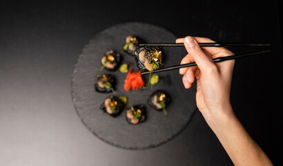 Hand with chopsticks holds a sushi. Custom sushi roll with black rice, crab meat, avocado, smoked...