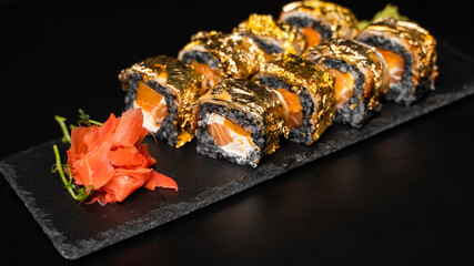 Custom sushi roll with nori, cuttlefish ink, fresh salmon, cream cheese, pepper pumpkin, eel, unagi sauce and edible gold leaf with red ginger, wasabi on a square plate on black table and background.