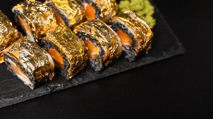 Custom sushi roll with nori, cuttlefish ink, fresh salmon, cream cheese, pepper pumpkin, eel, unagi sauce and edible gold leaf with red ginger, wasabi on a square plate on black table and background.