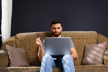 A young man in home clothes is sitting at home on a comfortable sofa with a laptop. Works and holds a bank card. Work online. Transfer of earned money.