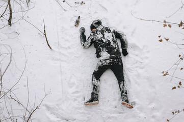 A drunken homeless and dirty man in a black coat lies, sleeps on his stomach on white snow in a...
