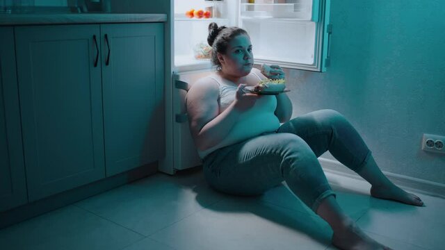 Young obese female eating sandwich, sitting near the fridge at night, calories