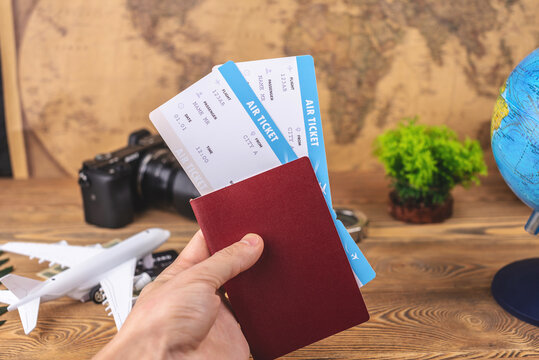 Hand is holding a passport and plane tickets over a wooden table with travel supplies. Vacation and booking concept