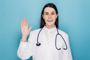Beautiful friendly young doctor woman wearing medical uniform waving saying hello happy and smiling, welcome gesture, isolated on blue studio background. Covid 19, virus, health and medicine concept
