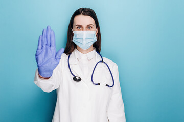 Serious woman doctor in medical face mask, with stethoscope and white coat, show stop sign,...