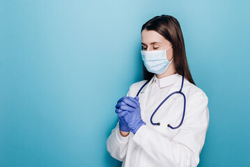 Young woman doctor in white medical gown sterile face mask gloves holding hands folded in prayer,...