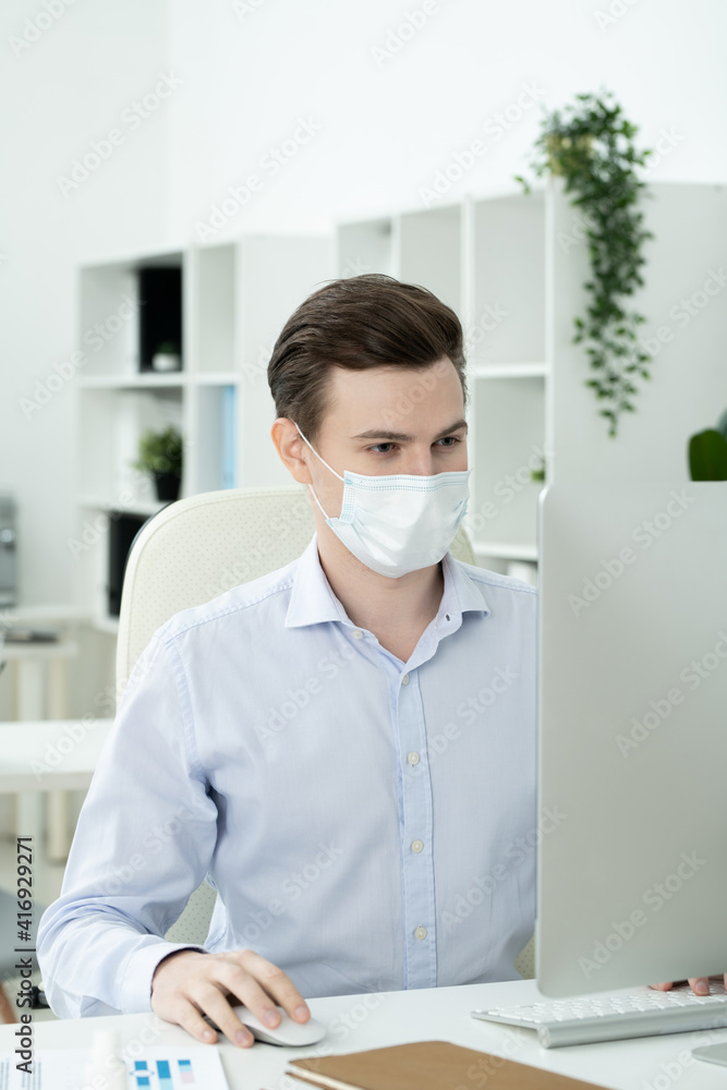 Poster young serious designer with protective mask on face looking at computer screen