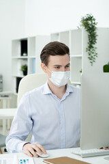 Young serious designer with protective mask on face looking at computer screen