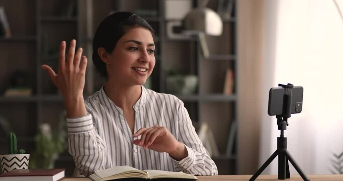 Happy young indian ethnicity female business trainer influencer blogger sharing professional news, recording educational business video on smartphone web camera, standing on stabilizer on table.