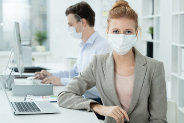 Young elegant businesswoman in protective mask looking at you against colleague