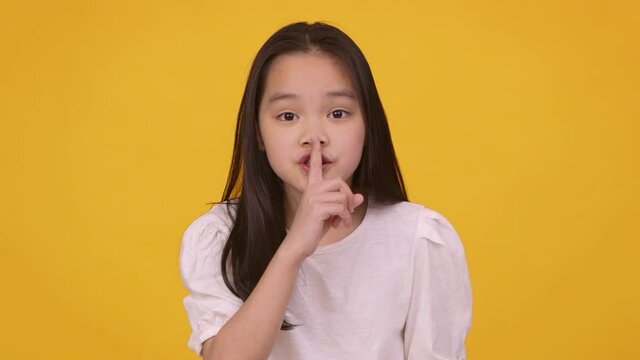 Keep our secret. Cute little asian girl putting finger on lips, showing hush gesture at camera, orange studio background