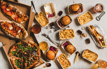 Lockdown fast food dinner from delivery service concept. Flat-lay of quarantine home party with burgers, french fries, sandwiches, pizza, beer in glasses over white tablecloth background, top view