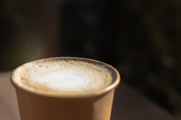 brown paper cup with cappuccino coffee with white foam on a wood table on a dark background