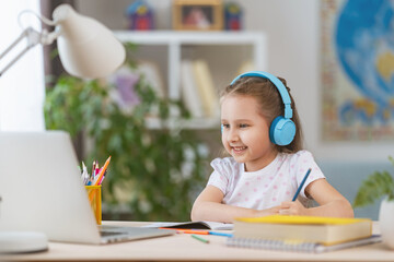 smart smiling little girl wearing headphones is learning online, using laptop remote learning. child listens to teacher and writes down task in notebook. interactive E-Education Distance Home learning