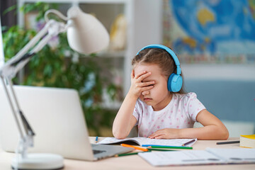 sad little girl, schoolgirl wearing headphones, uses laptop to study at home. child is bored to...