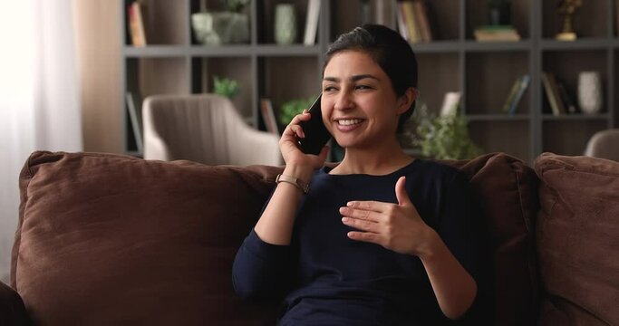 Pleasant smiling young indian mixed race woman resting on comfortable couch, speaking with friends, communicating distantly by smartphone conversation, sharing or discussing life news at home.