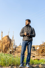 Naklejka na ściany i meble An indian handsome man standing on a farm wearing black coat with a camera in his hand. freedom and travel concept.
