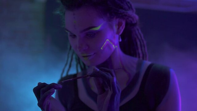 Making up a Girl with Dreadlocks in Neon UF Light. Model Girl with Fluorescent Creative Psychedelic MakeUp, Art Design of Female Disco Dancer Model in UV, Colorful Abstract Make-Up. Dancing Lady