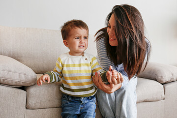 Portrait of adorable little boy sitting on the textile couch with his mom and crying. Upset toddler...