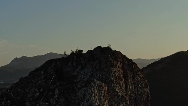 Hiker in Baseball Cap with Backpack and Nordic Walking Sticks Climbs a Rocky Top of a Mountain on the Background of a Mountain Landscape and Stands in the Rays of Sunset. Aerial view. Surveying by dro