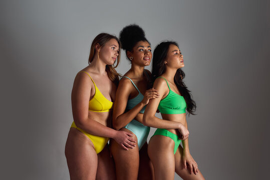 Three confident young women with different body shapes wearing colorful underwear looking aside, posing together isolated over grey background