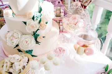 Candy bar. White wedding cake decorated by flowers standing of festive table with deserts and cupcakes