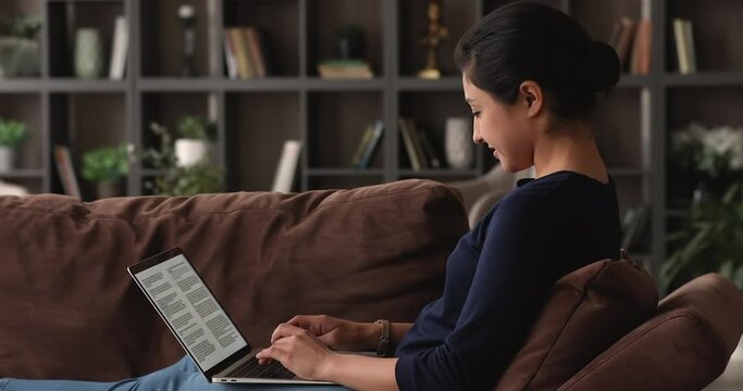 Side view happy smart young mixed race indian woman relaxing on comfortable couch, working with electronic document on computer, editing text, writing novel or preparing scientific article at home.