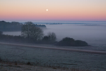 an early morning mist filled valley with the snow moon low in the sunrise sky 