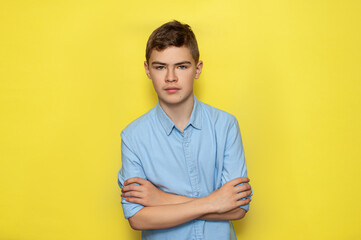 A teenage Caucasian boy in a blue shirt on a yellow background folded his arms on his chest. Serious schoolboy.