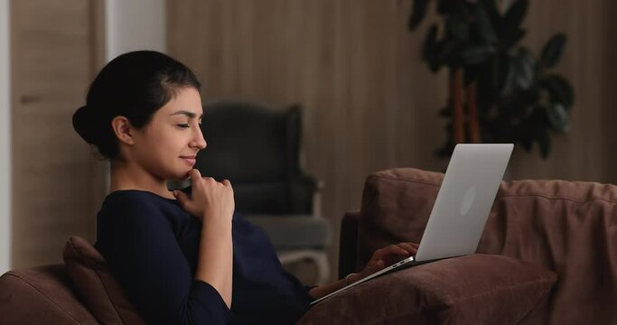 Smiling millennial indian ethnicity woman sitting on sofa with computer in pillow on laps, typing message in social network, communicating distantly, creating new ideas or web surfing information.