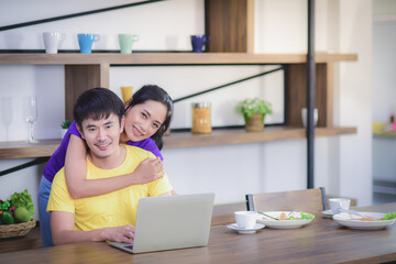 Asian couples are eating breakfast and coffee with a white coffee cup In dining room happily The husband uses a laptop to track news on social media and works online and the wife embraces from behind.
