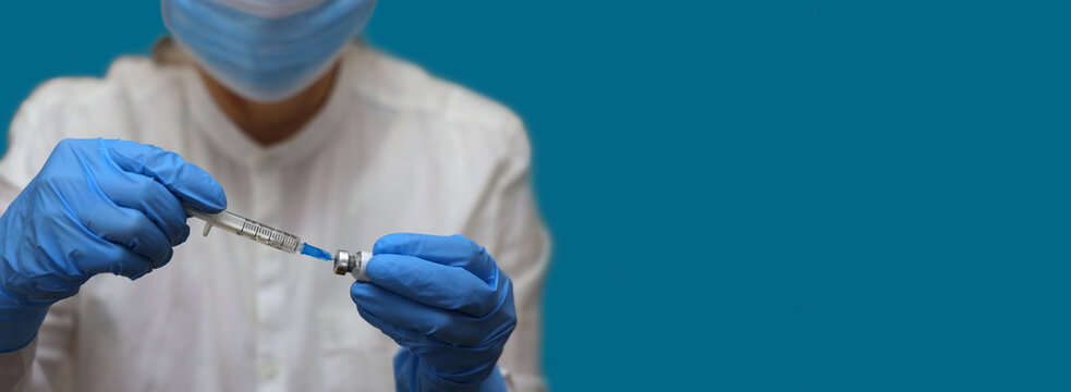Nurse wearing in medical gloves and face mask fills syringe from vial of COVID vaccine for revaccination population against omicron mutation. Close-up on blue background with copy space.