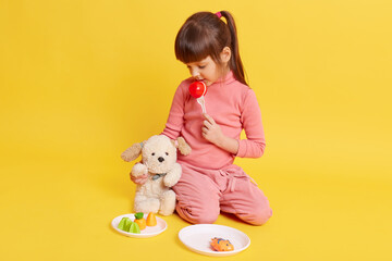 Small cute girl playing with soft dog and plastic vegetables, kid imaging eating food, puts dog next to her and looking at it, wearing pink attire has ponytail, isolated over yellow background.