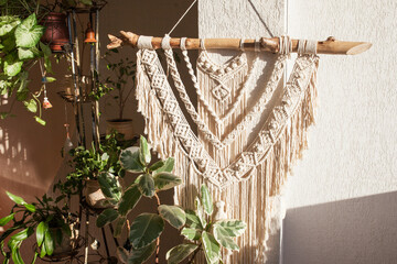 Handmade macrame  100% cotton wall hanging with green leafes on a white wall.  Modern eco macrame decoration with wooden stick in the interior. Bright solar light