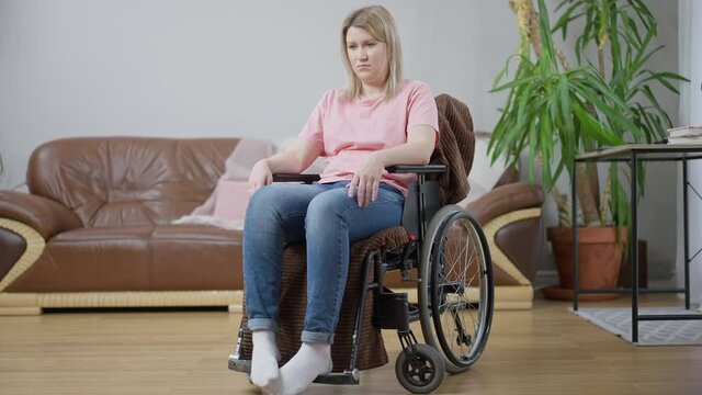 Wide shot portrait of depressed frustrated disabled woman sitting in wheelchair at home thinking. Young paralyzed Caucasian invalid alone indoors. Frustration and disability concept.