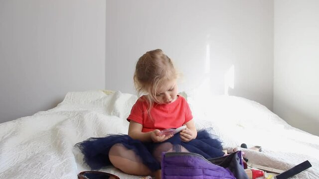 A child girl sits on the bed and paints her cheeks with a brush.