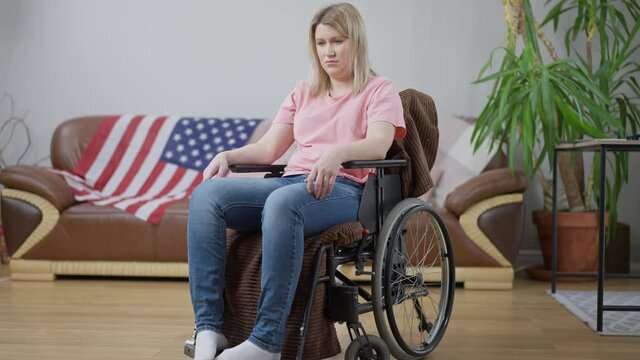Young frustrated American woman sitting in wheelchair in living room at home with national flag on couch at background. Portrait of depressed disabled person indoors. Handicap concept.