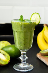 Very tasty green cocktail for detoxification. Delicious green smoothie and fresh fruits on black table. Healthy green smoothie with banana, spinach, avocado, apple and mint in a glass. Vegan food.