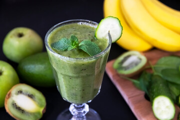 Very tasty green cocktail for detoxification. Delicious green smoothie and fresh fruits on black table. Healthy green smoothie with banana, spinach, avocado, apple and mint in a glass. Vegan food.