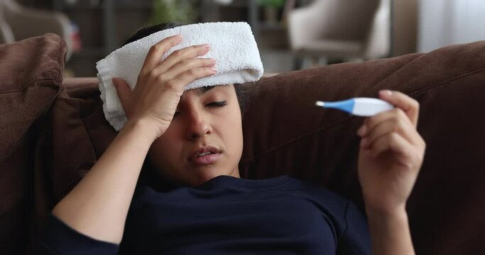 Unhealthy young indian ethnicity woman making head compress with wet towel, suffering from headache, measuring body temperature with electronic thermometer, having first flu grippe covid19 symptoms.