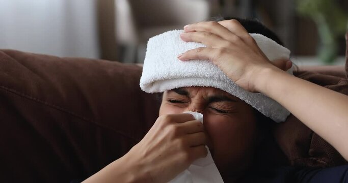Close up head shot unhealthy young indian woman with applied on forehead cold towel compress sneezing in paper tissue, suffering from runny nose, caught cold, feeling first grippe flu symptoms.