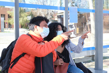 Young latin couple wearing protective face mask and carrying guitar case pointing at something, sitting at bus stop. New normal in public transport.