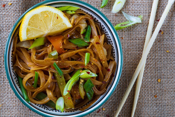 Stir-fried rice noodles with chicken and vegetables (Carrot, bell pepper, onion, lemon, chili) dish in bowl. Gluten free traditional Asian cuisine with chopsticks. Top view, closeup, rustic background