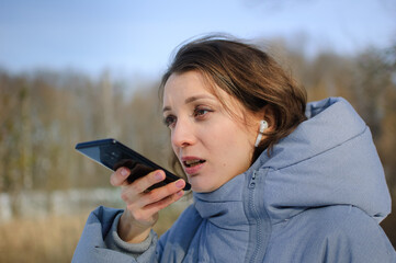 Lady in blue coat is sending audio voice message explaining something on smart phone at outdoor talking to mobile assistant. Girl using smartphone voice recognition and dictation