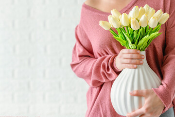 Woman in a pink pullover holding vase with white tulips flowers opposite brick wall. Copy space