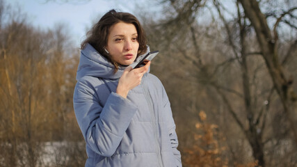 Lady in blue coat is sending audio voice message explaining something on smart phone at outdoor...
