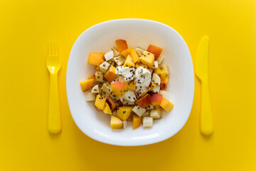Healthy nutrition fruit salad with soft cheese and flax seeds in white plate on yellow background with yellow knife and fork. Dieting and balance food.