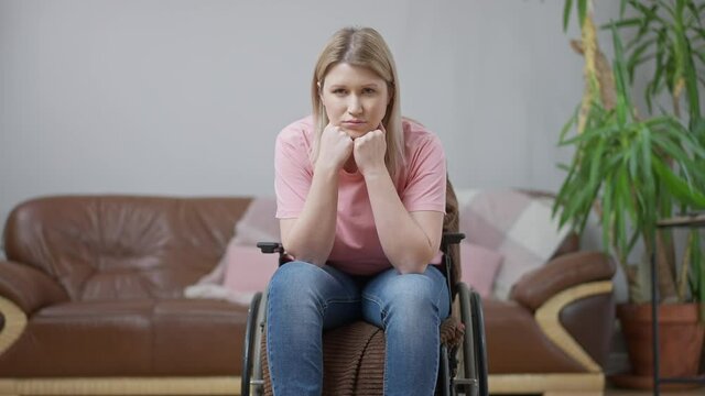 Unhappy paralyzed young Caucasian woman sitting in wheelchair in living room looking at camera. Portrait of sad paraplegic lady posing at home indoors. Disability and lifestyle concept.