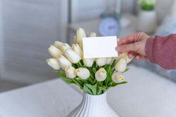 Woman pulling blank greeting card from bouquet of white tulips flowers. Copy space. Mock up